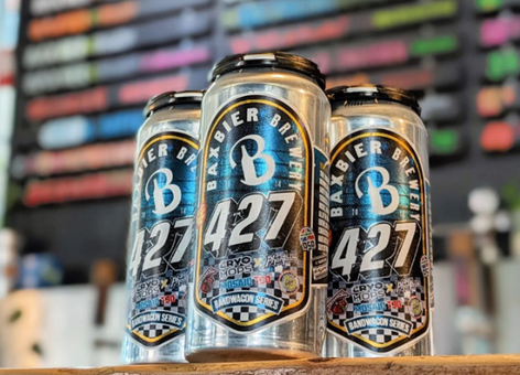 Four Baxbier Brewery 427 IPA cans on a wooden counter with a colorful, blurred tap list in the background.