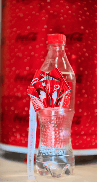 Een doorzichtige plastic fles met een rode Coca-Cola-strik en -lint, afgebeeld tegen een felrode, feestelijke Coca-Cola-achtergrond.