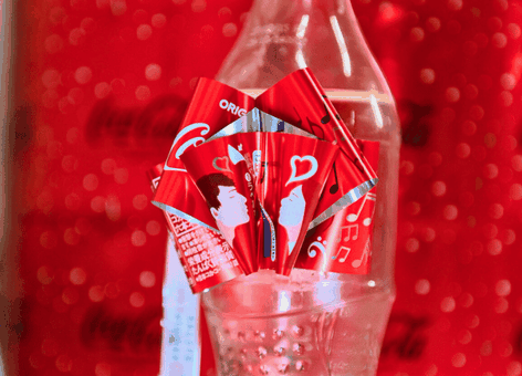 Een doorzichtige plastic fles met een rode Coca-Cola-strik en -lint, afgebeeld tegen een felrode, feestelijke Coca-Cola-achtergrond.