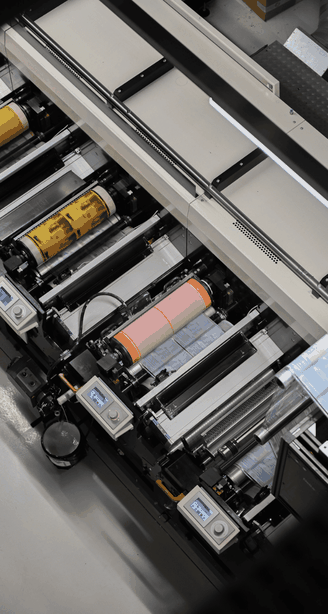 Top view of an industrial printing press with colored cylinders and digital control panels in operation.