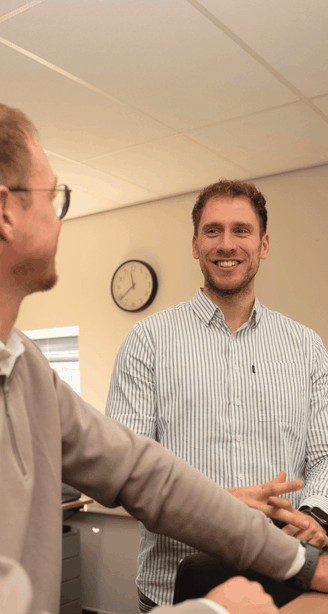 Two colleagues chat and smile in an office, with a wall clock and desks visible in the background.