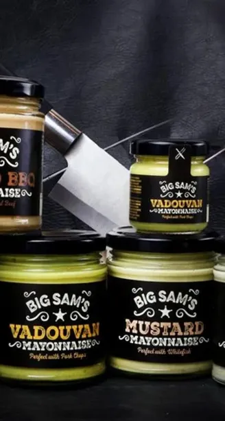 Assorted jars of Big Sam’s flavored mayonnaise with knives in the background on a dark surface.
