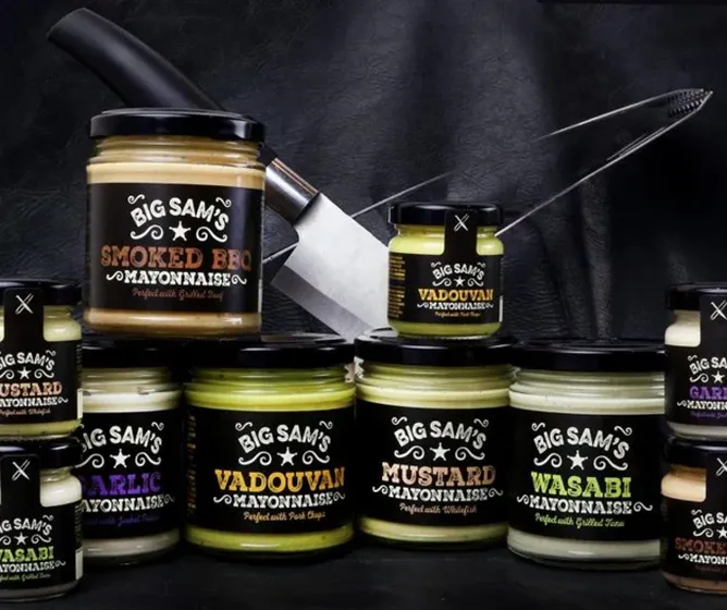 Assorted jars of Big Sam’s flavored mayonnaise with knives in the background on a dark surface.