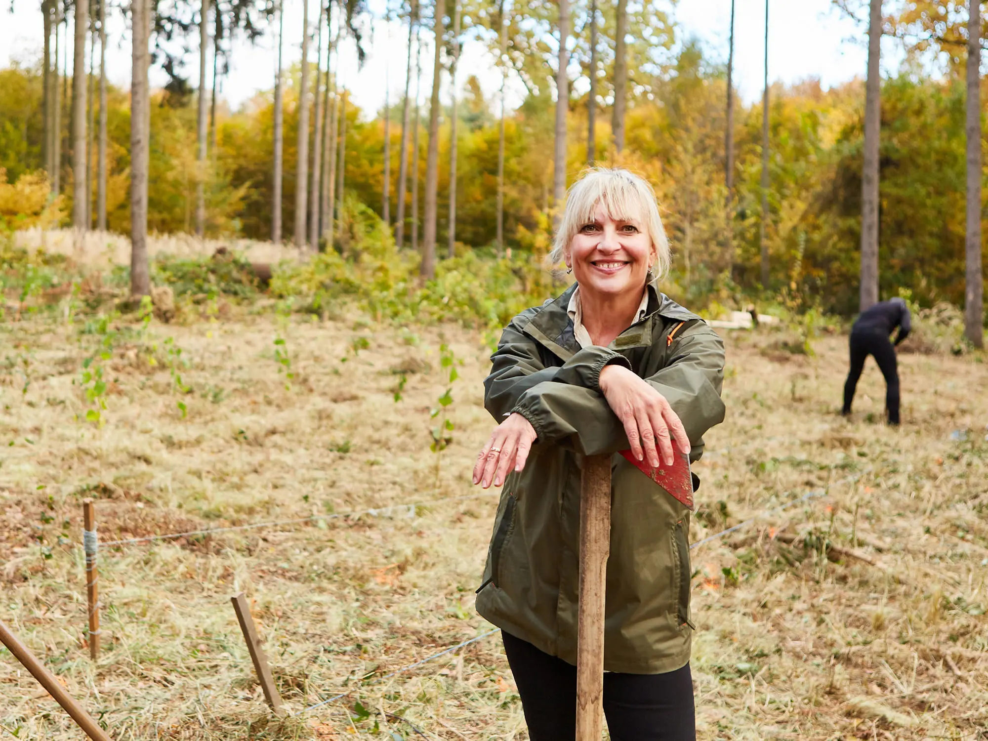 Eine Frau in grüner Jacke lehnt lächelnd auf einer Waldlichtung an einer Schaufel. Im Hintergrund arbeitet eine weitere Person. Die Bäume haben herbstliches Laub.