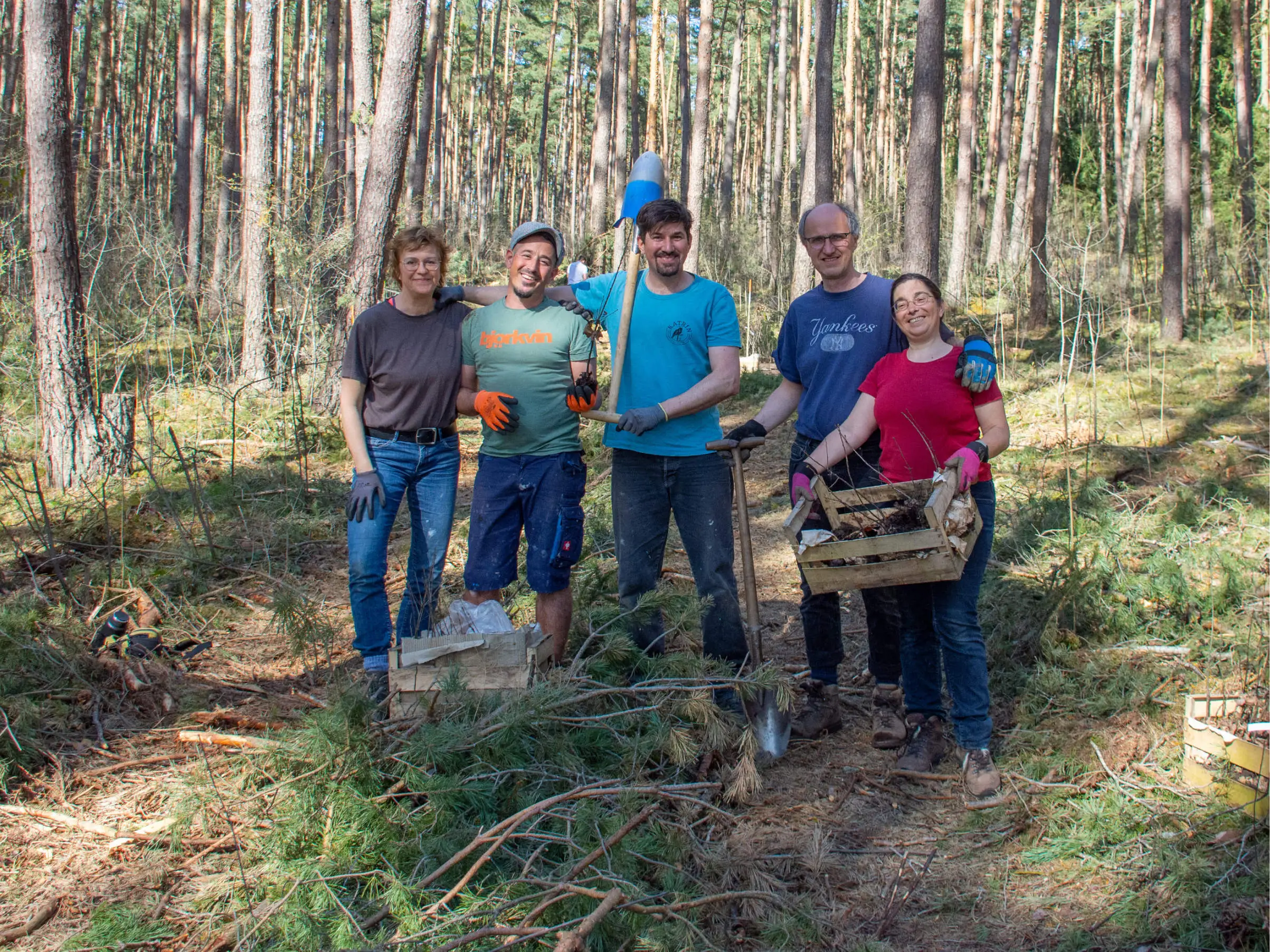 Fünf lächelnde Menschen posieren in einem Wald, halten Werkzeuge und eine Kiste mit Setzlingen in den Händen und deuten damit eine Baumpflanz- oder Naturschutzaktion an.