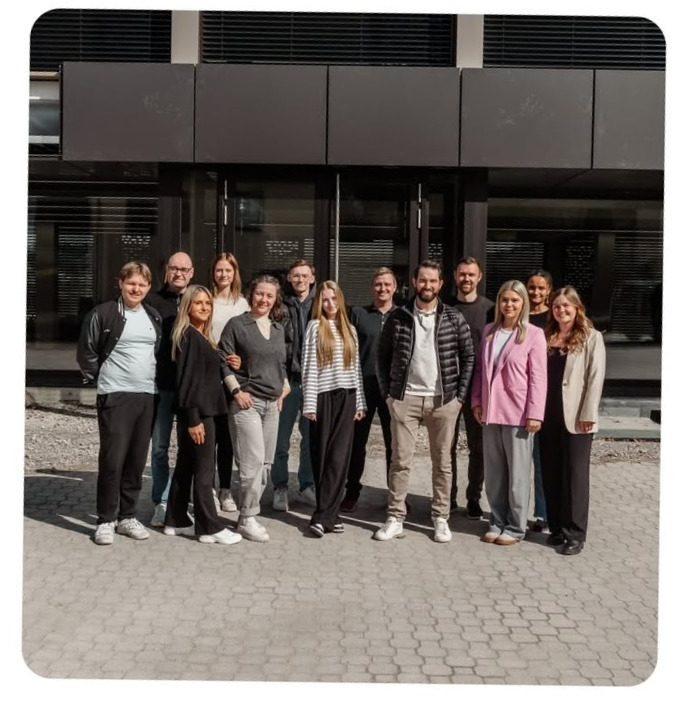 Eine Gruppe von dreizehn Personen des Unternehmens Hosta steht an einem sonnigen Tag vor einem modernen Gebäude, lächelt und posiert für ein Foto.