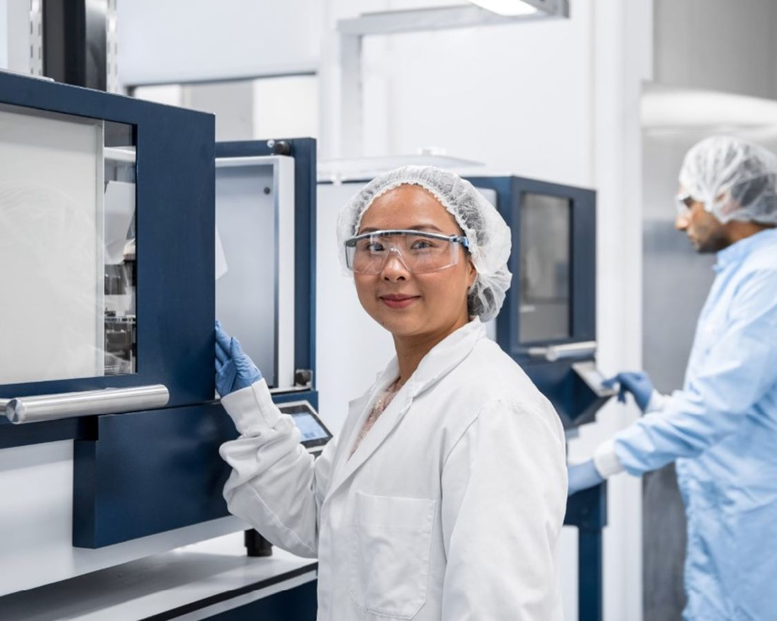 A female scientist in a lab coat and safety glasses smiling while operating equipment in a laboratory, with a male colleague in the background, showcasing the innovative research at CAM Bioceramics.