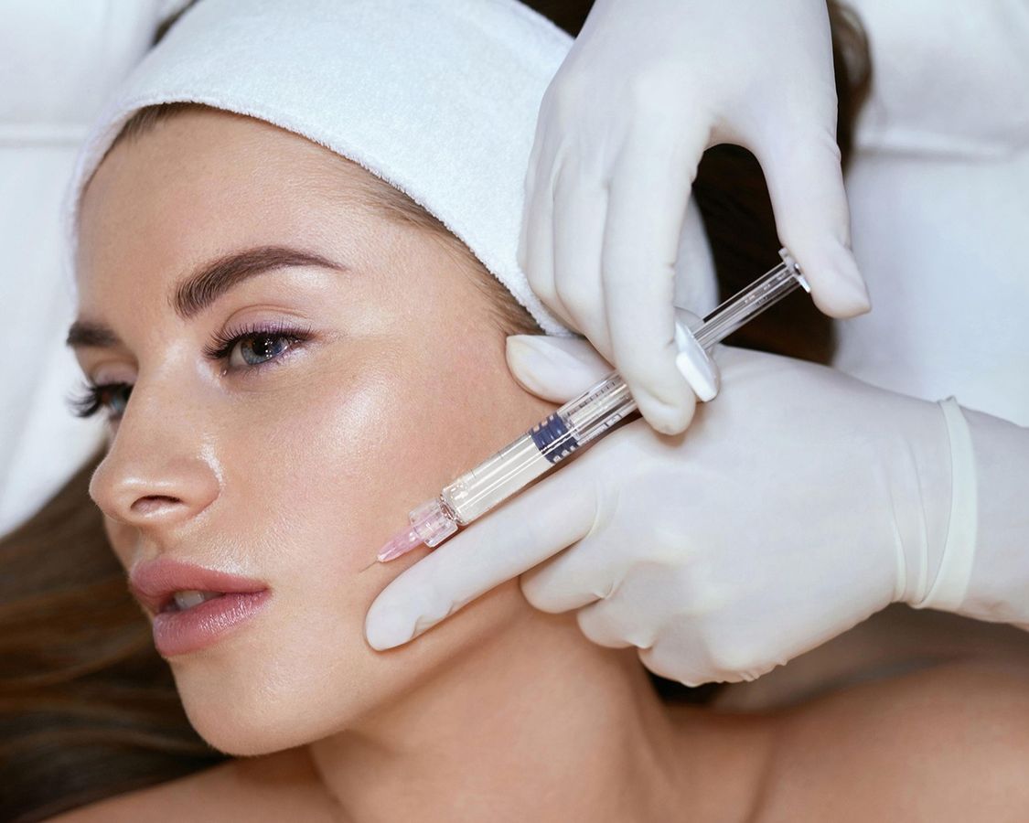 A close-up image of a woman receiving a cosmetic injection on her cheek. She has long, flowing hair and is wearing a white headband. A gloved hand administers the injection with a syringe, emphasizing a professional skincare treatment. The background is softly blurred, focusing on the procedure.