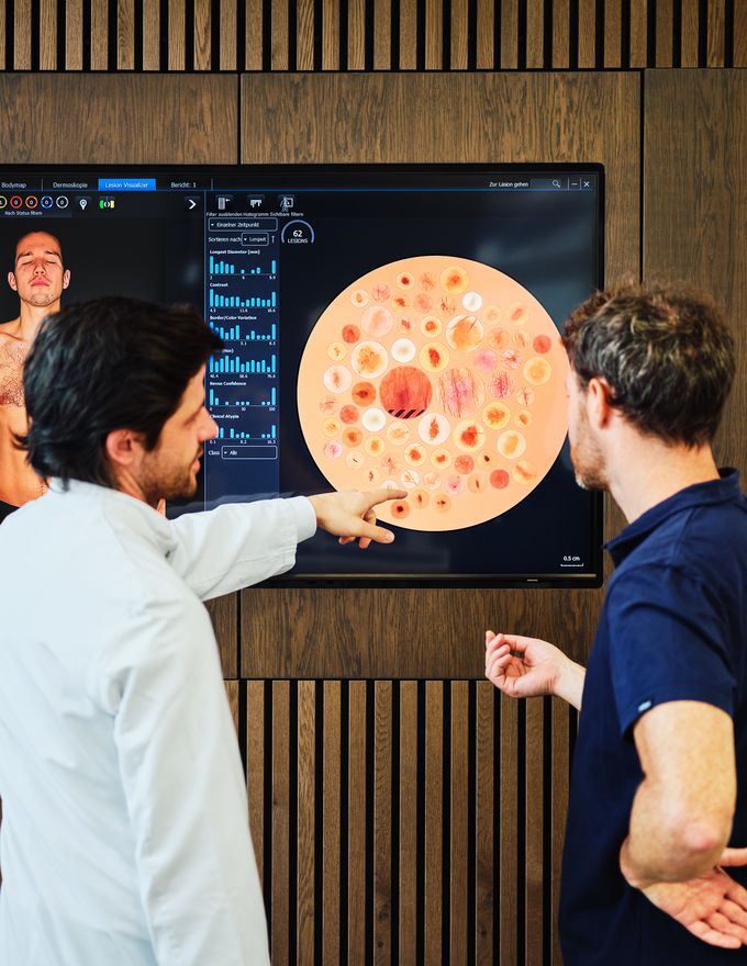 Two men analyzing a large screen displaying a medical image and data, with one pointing at the screen, in a modern office setting executing remote digital skin cancer screenings.
