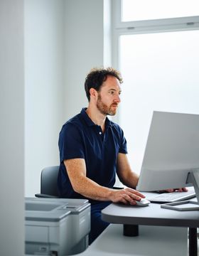 A man in a blue shirt works at a computer in a bright office, with a printer nearby performing digital remote skin cancer screeing for patients.