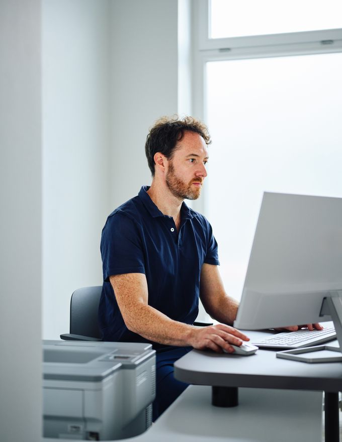 A man in a blue shirt works at a computer in a bright office, with a printer nearby performing digital remote skin cancer screeing for patients.