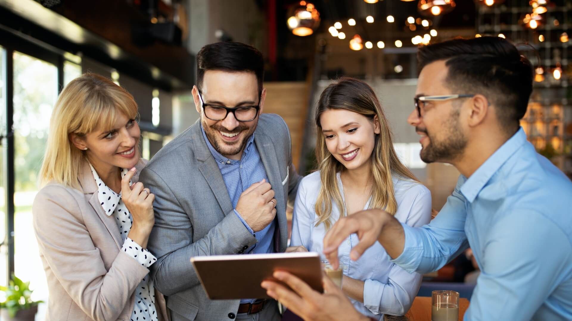 Four people smiling and looking at a tablet in a modern office setting with warm lighting.