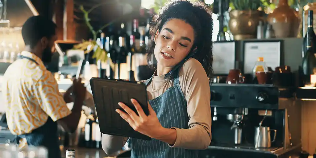 Camarera con delantal atendiendo una llamada mientras gestiona reservas desde una tablet en la barra de un restaurante.