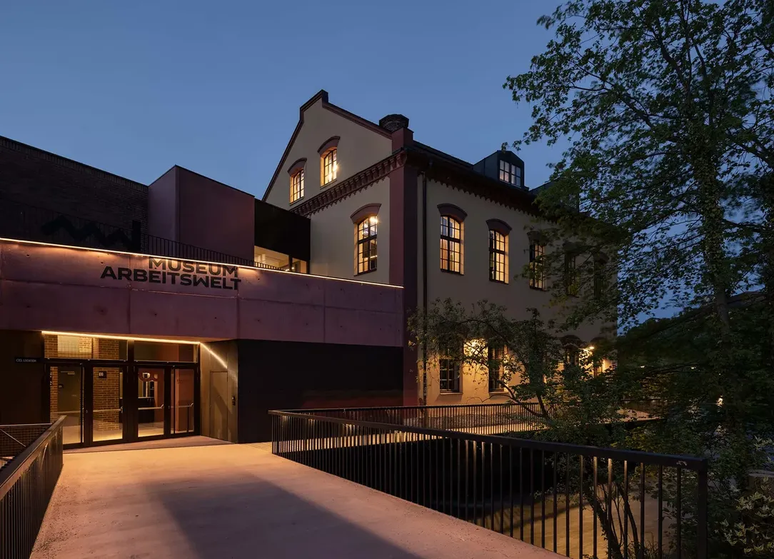 Exterior view of a lit building at dusk with "Museum Arbeitswelt" signage, featuring modern and traditional architectural elements and surrounding trees.