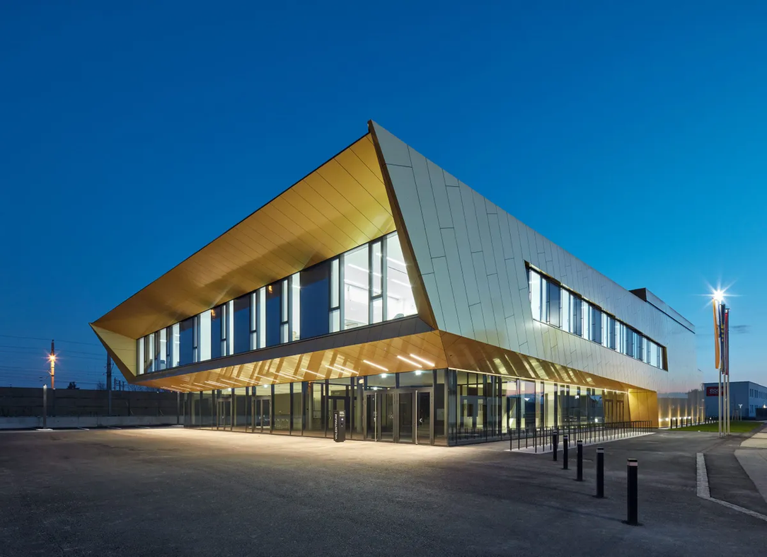 Modern building with a distinctive geometric design and large windows, illuminated at dusk against a clear blue sky.