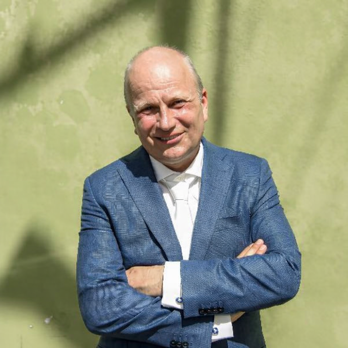 Man in blauw pak voor een groene muur staat met armen over elkaar en lacht naar de camera