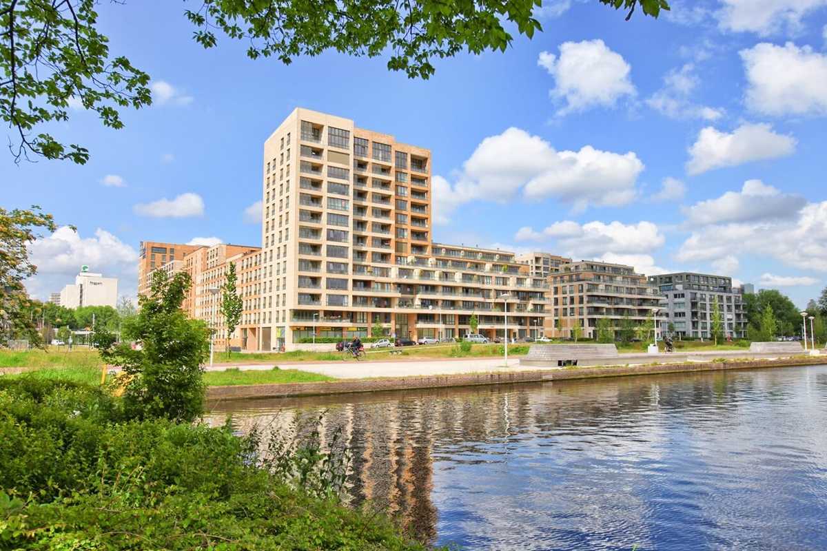 Complex van huurwoningen in Amsterdam. Het gebouw heeft verschillende beige en oranjetinten en staat langs het water. Op de achtergrond zien we blauwe lucht en bomen.