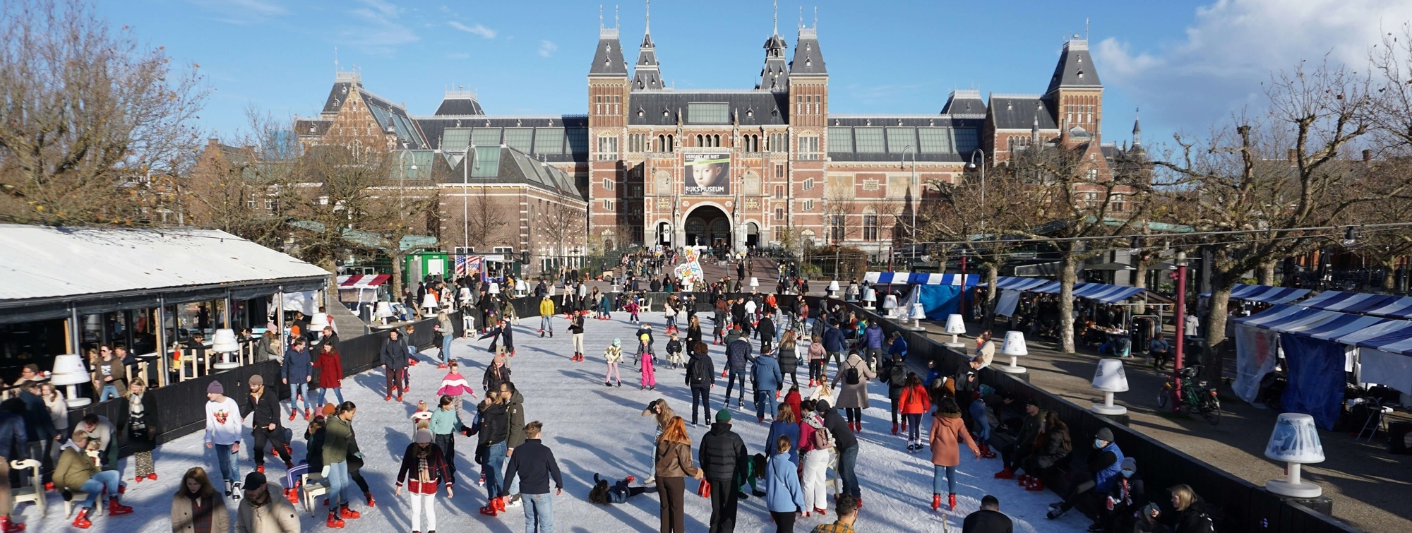 foto van de schaatsbaan op het museumplein voor het imposante rijksmuseum in Amsterdam. de zon schijnt en er zijn veel mensen op schaatsen