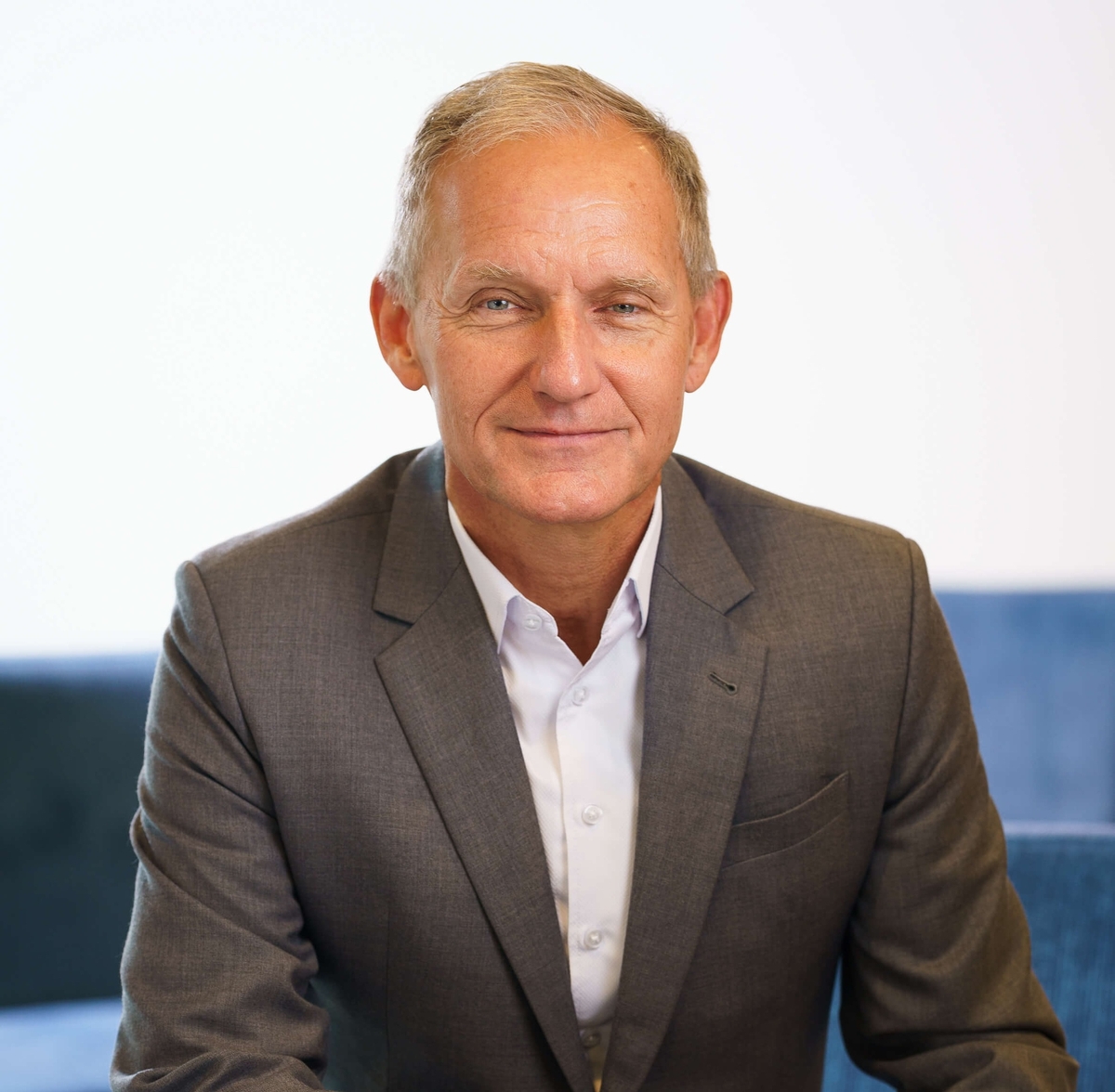 portret van een blanke man in een grijs pak met witte blouse die zit op een blauwe vorm