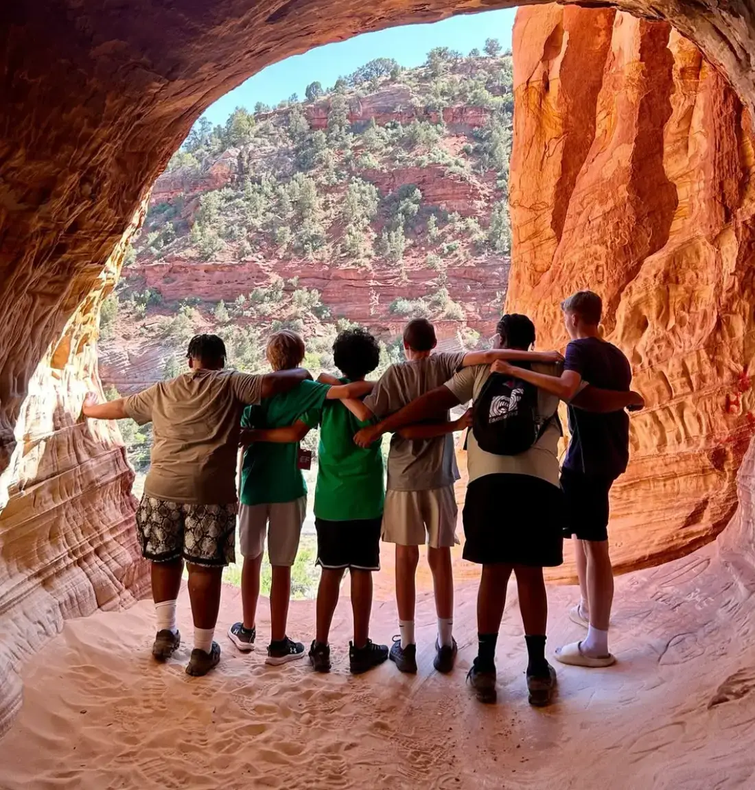 Students standing arm in arm inside a sandstone cave overlooking red rock cliffs.