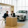 Verkoper overloopt een offerte met een klant aan een tafel in de showroom, naast een witte SUV, om salonvoorwaarden te vergelijken