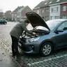 Man buigt zich onder de open motorkap van een grijze auto die niet start, geparkeerd langs een straat met kasseien en rijhuizen in de herfst