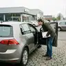 Man met een klembord buigt zich door het open portier van een grijze hatchback om het interieur te controleren voor een proefrit, op de parking van een autodealer