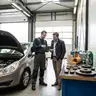 Mechanieker overloopt op een tablet de kosten van een groot onderhoud met een klant in de werkplaats, naast een auto met open motorkap en een werkbank met remschijven, remblokken, filters en olie