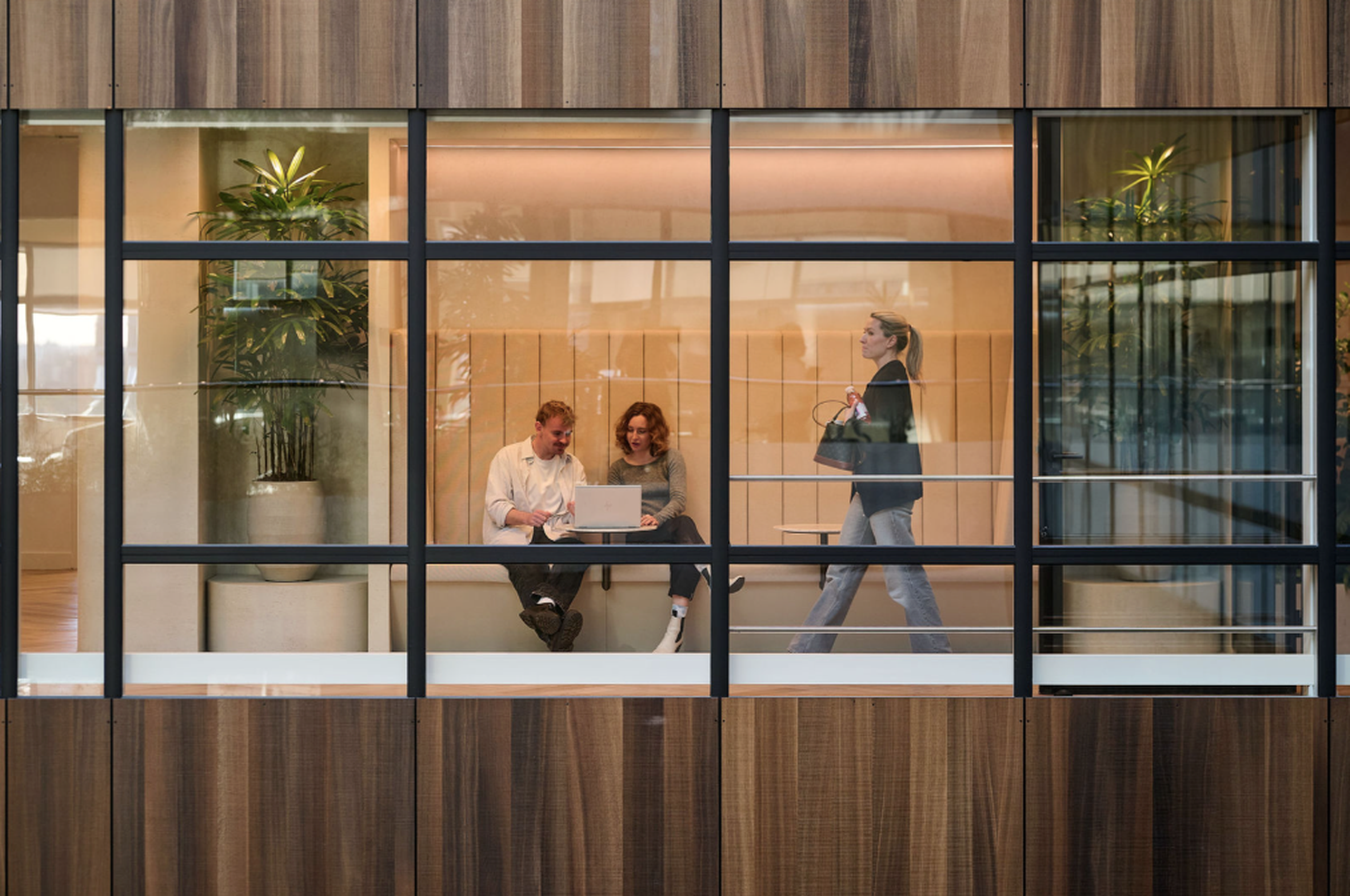 Two people collaborate on a laptop inside a modern office with plants, while a woman walks by outside, through a wooden and glass facade.