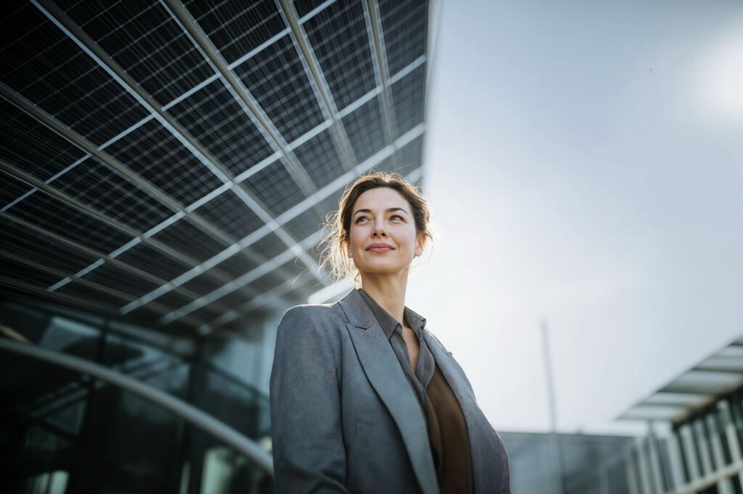 Smiling woman in a suit stands outdoors beneath solar panels, with a modern building in the background under a clear sky.
