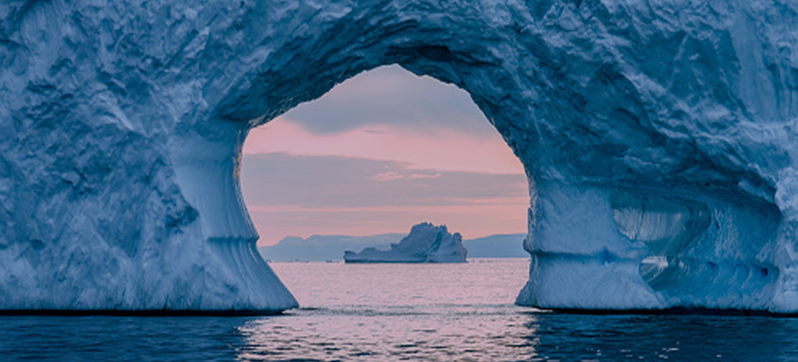 Iceberg with an arch frames a distant iceberg on calm water, under a pink sky at sunset.