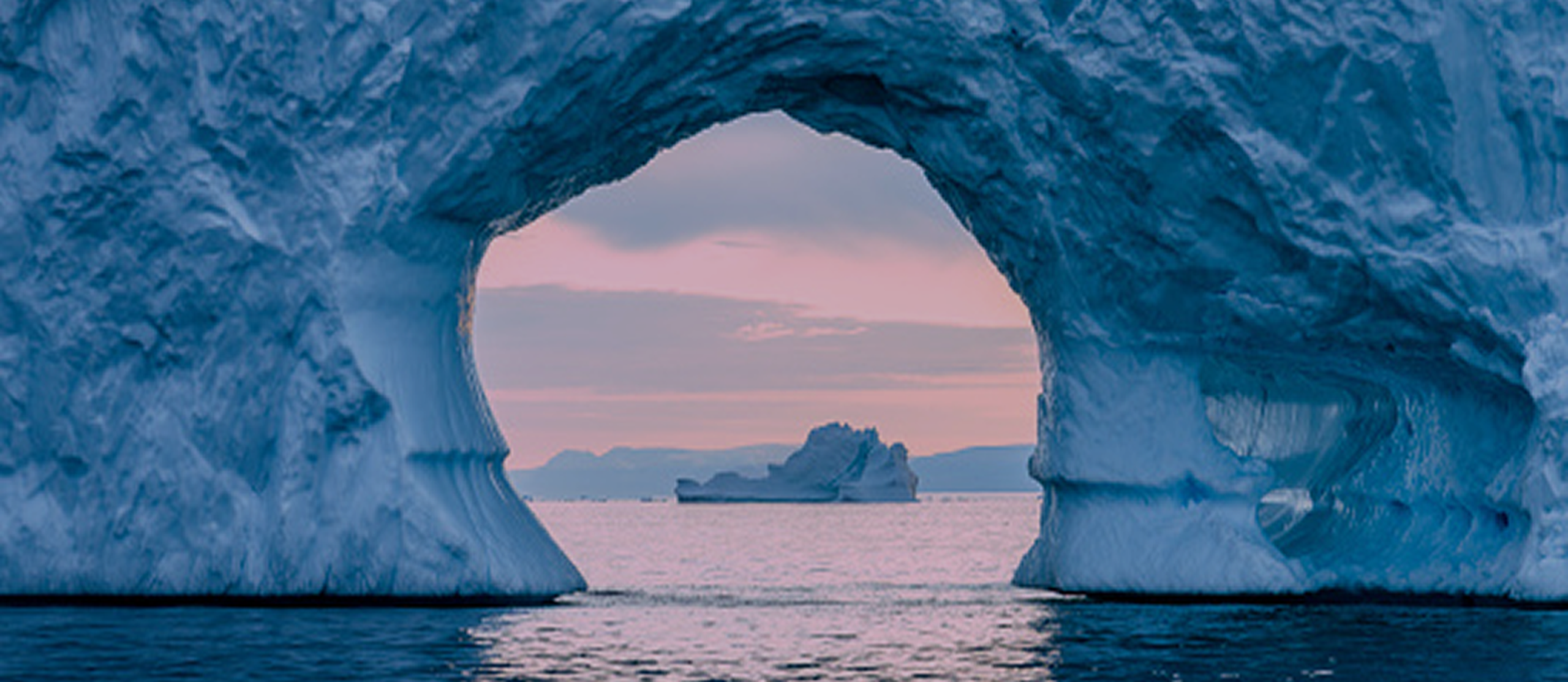 Iceberg with an arch frames a distant iceberg on calm water, under a pink sky at sunset.