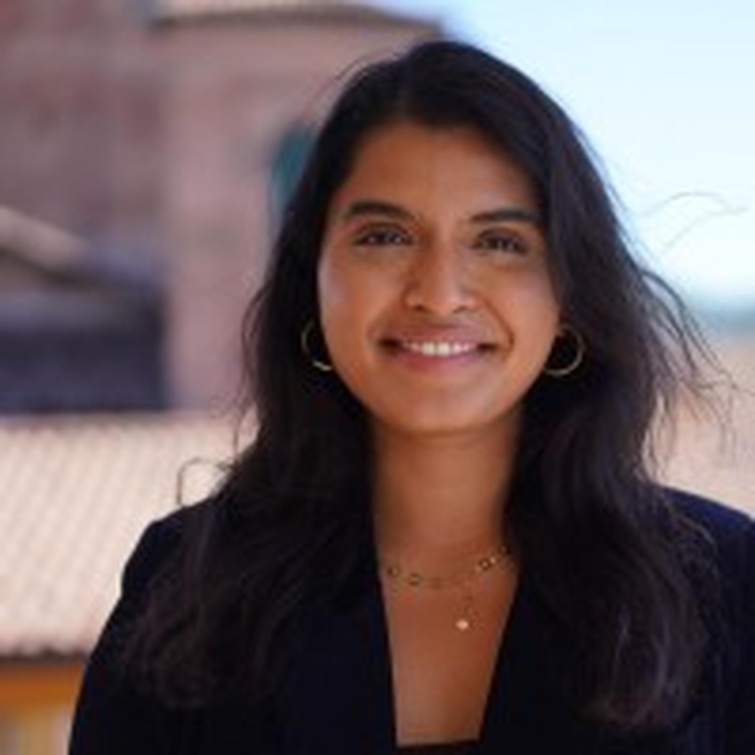A woman with long dark hair smiles at the camera; she is wearing hoop earrings and a necklace, with a blurry outdoor setting in the background.