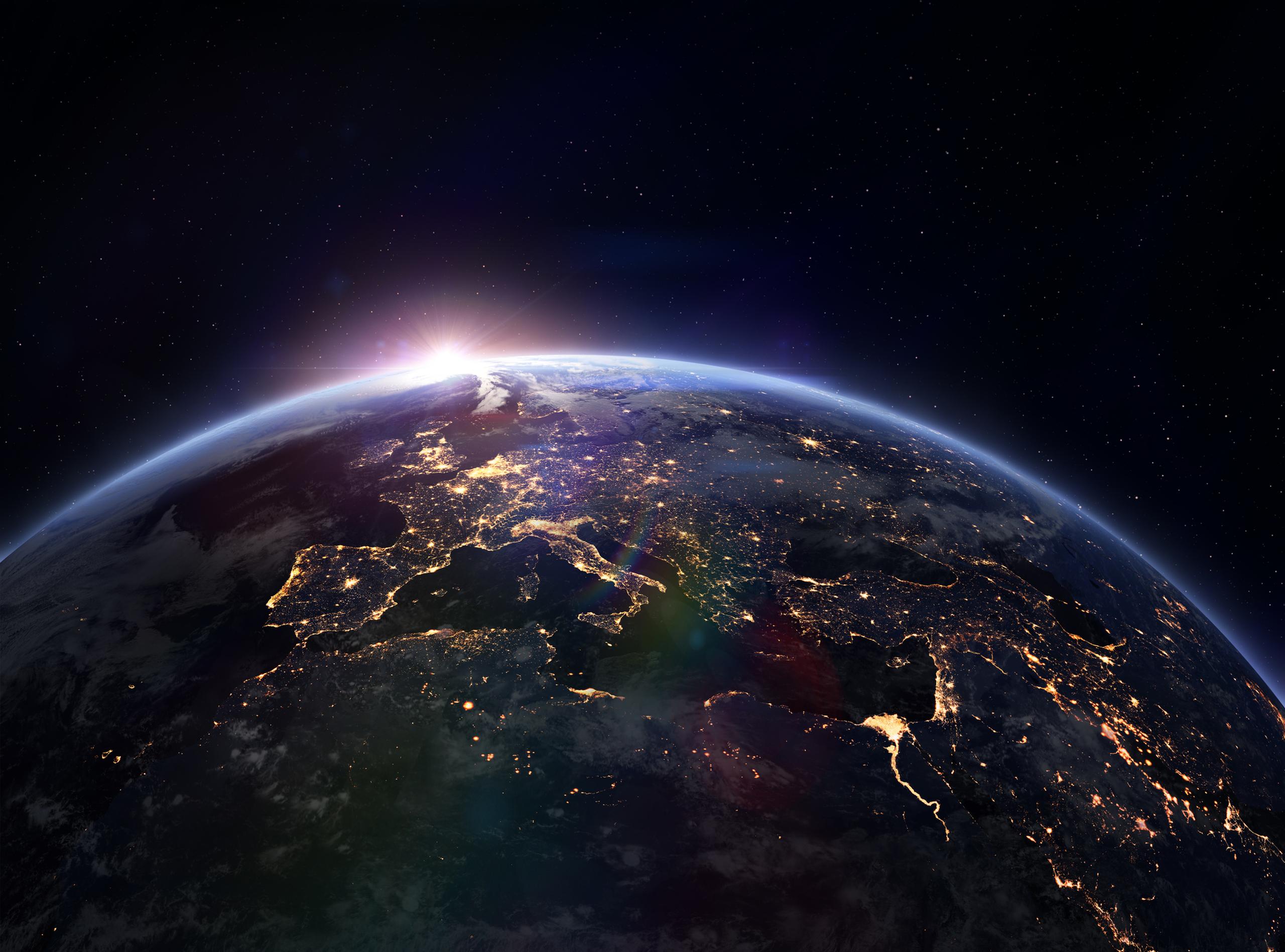 View of Earth from space at night, with city lights visible and a sunrise emerging over the horizon.