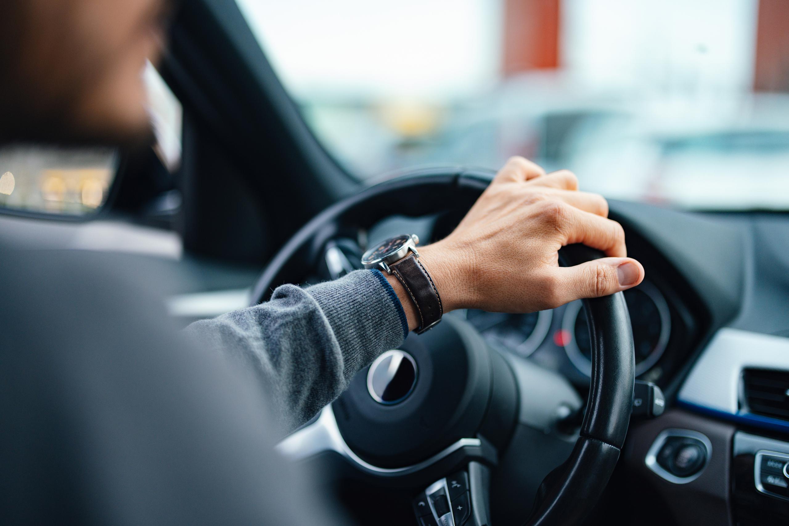 Close-up of a person in a sweater holding a car steering wheel, with a car dashboard visible.