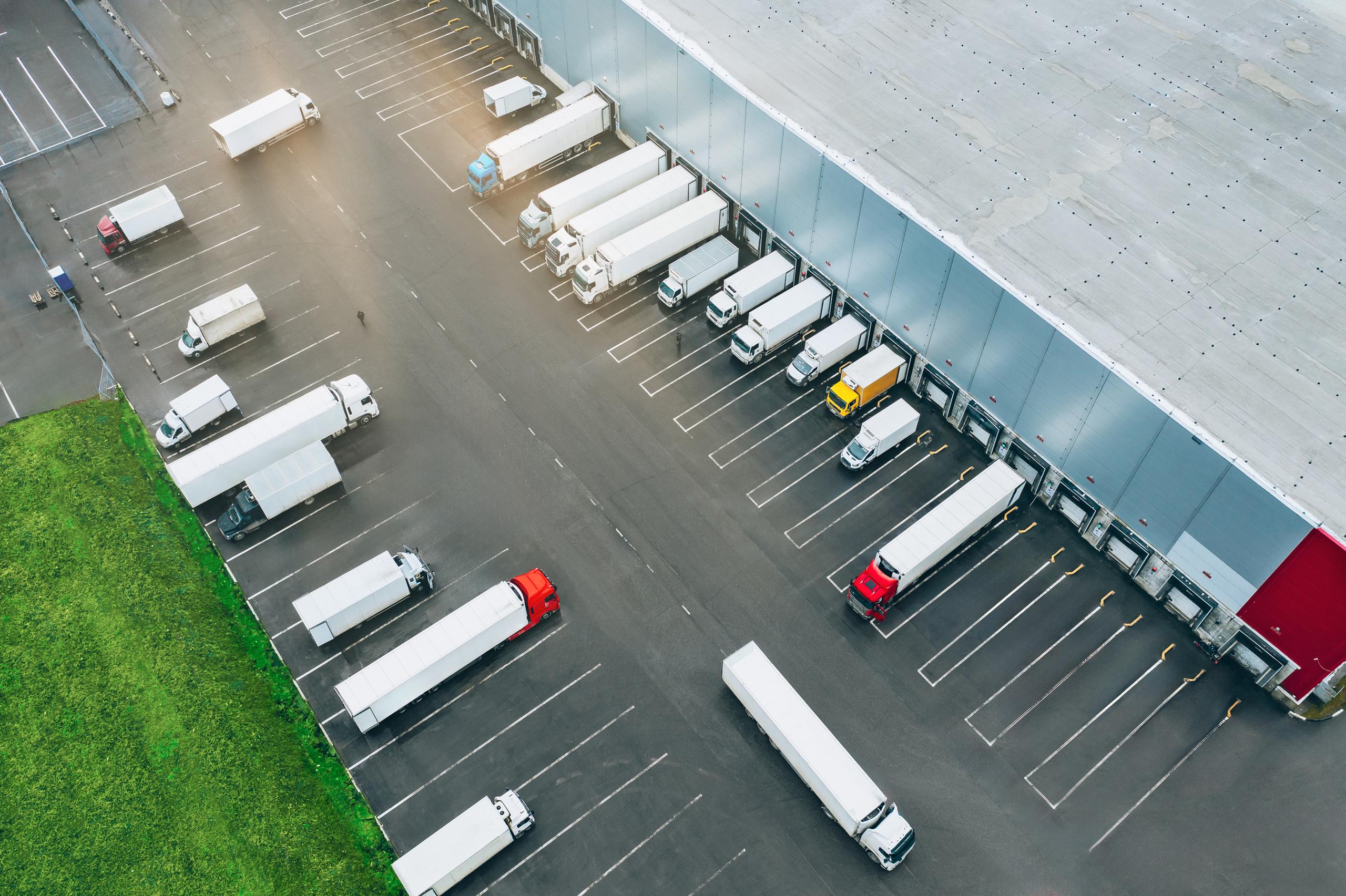 Aerial view of a distribution center with numerous trucks parked at loading bays and in a large parking lot, adjacent to a green grassy area.