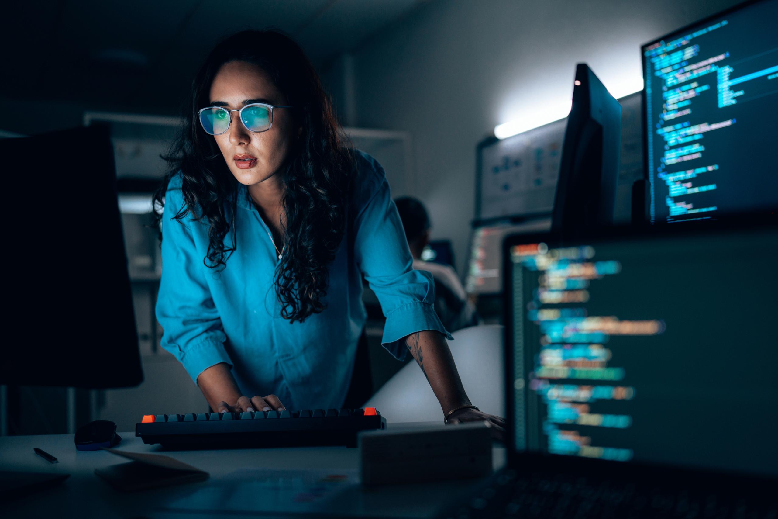 A woman wearing glasses, focused on a computer screen displaying code, works in a dimly lit office with multiple monitors.
