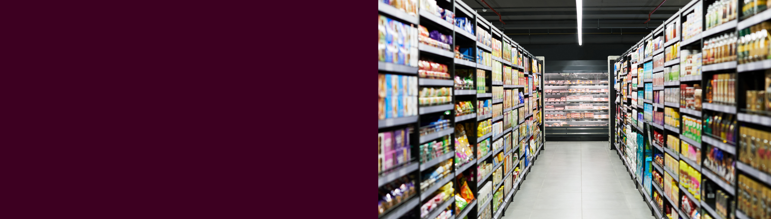 A well-lit grocery store aisle with fully stocked shelves on both sides, leading to refrigerated sections in the background.