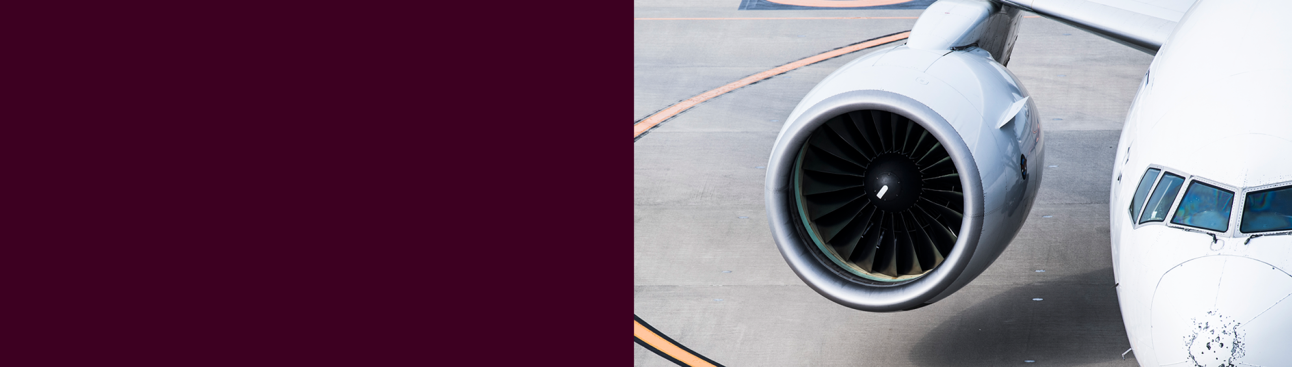 Close-up of a commercial airplane's cockpit and right engine on an airport tarmac with a maroon color block on the left.