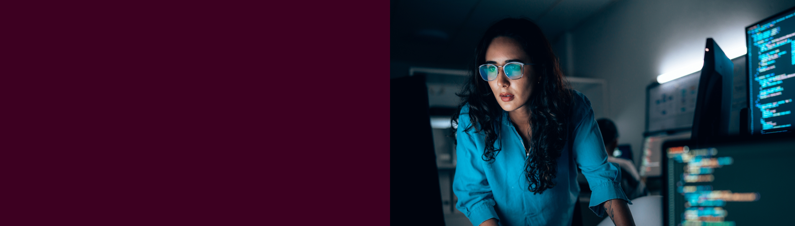 A woman in glasses focuses intently on a computer screen displaying code in a dimly lit office.