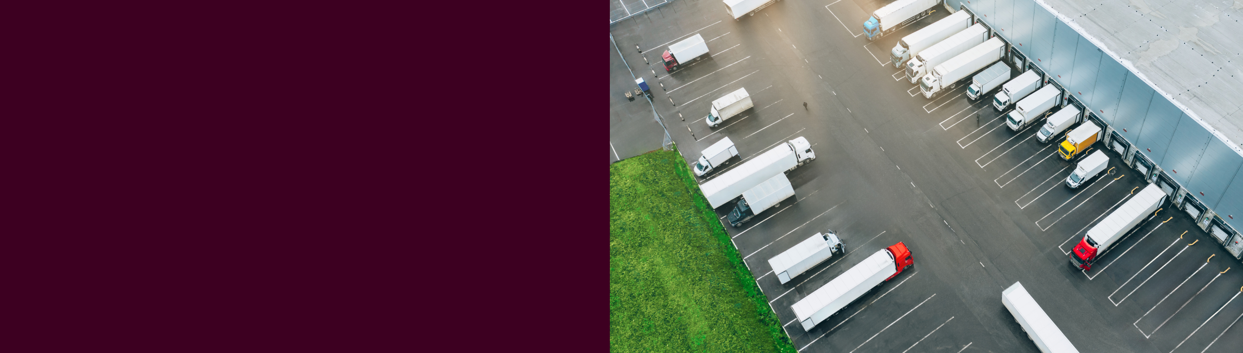 Aerial view of a warehouse parking lot with numerous trucks at loading docks, surrounded by a patch of green grass, with a deep maroon background on the left.
