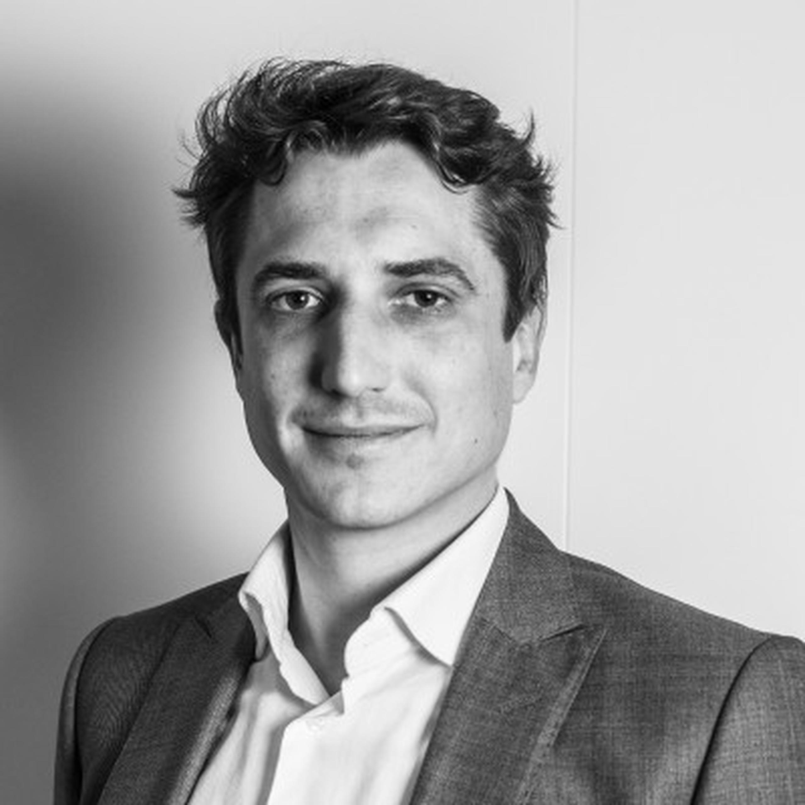 Man in a suit and white shirt smiling slightly, gazing at the camera. Black and white portrait.