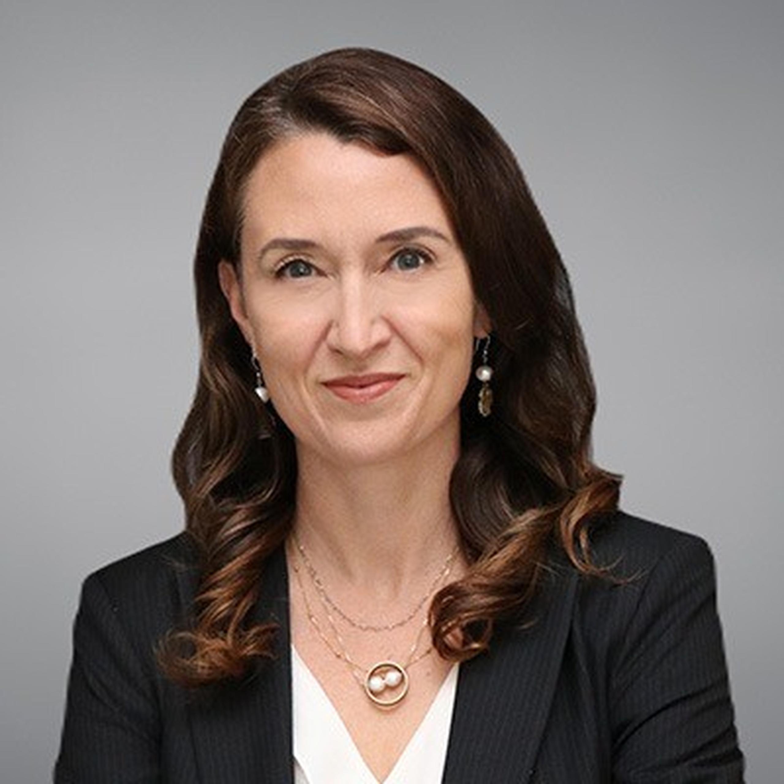 Woman with long brown hair, wearing a black blazer and white blouse, smiles slightly against a gray background.