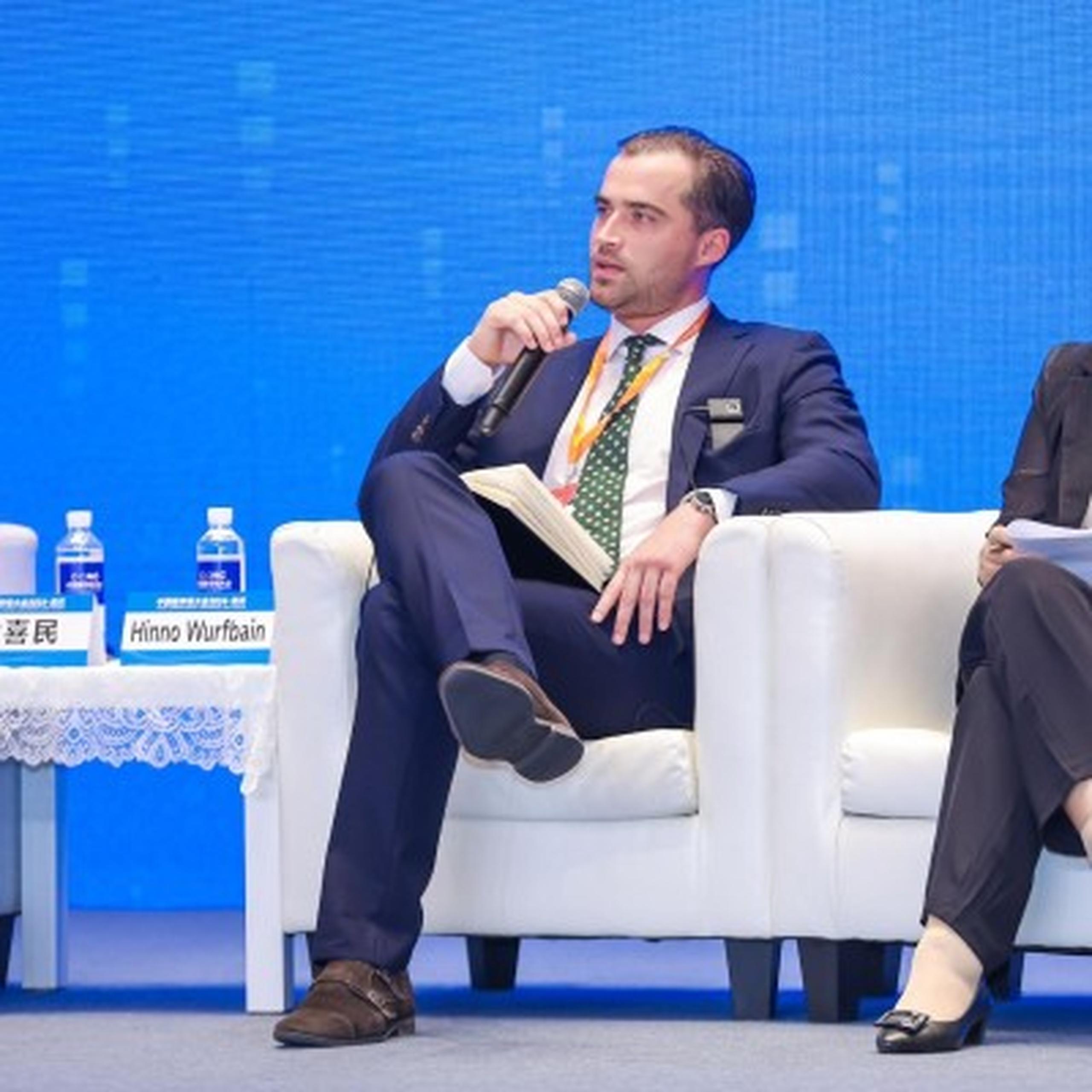Man in suit speaking into a microphone while seated on a panel with a blue backdrop and bottled water visible.