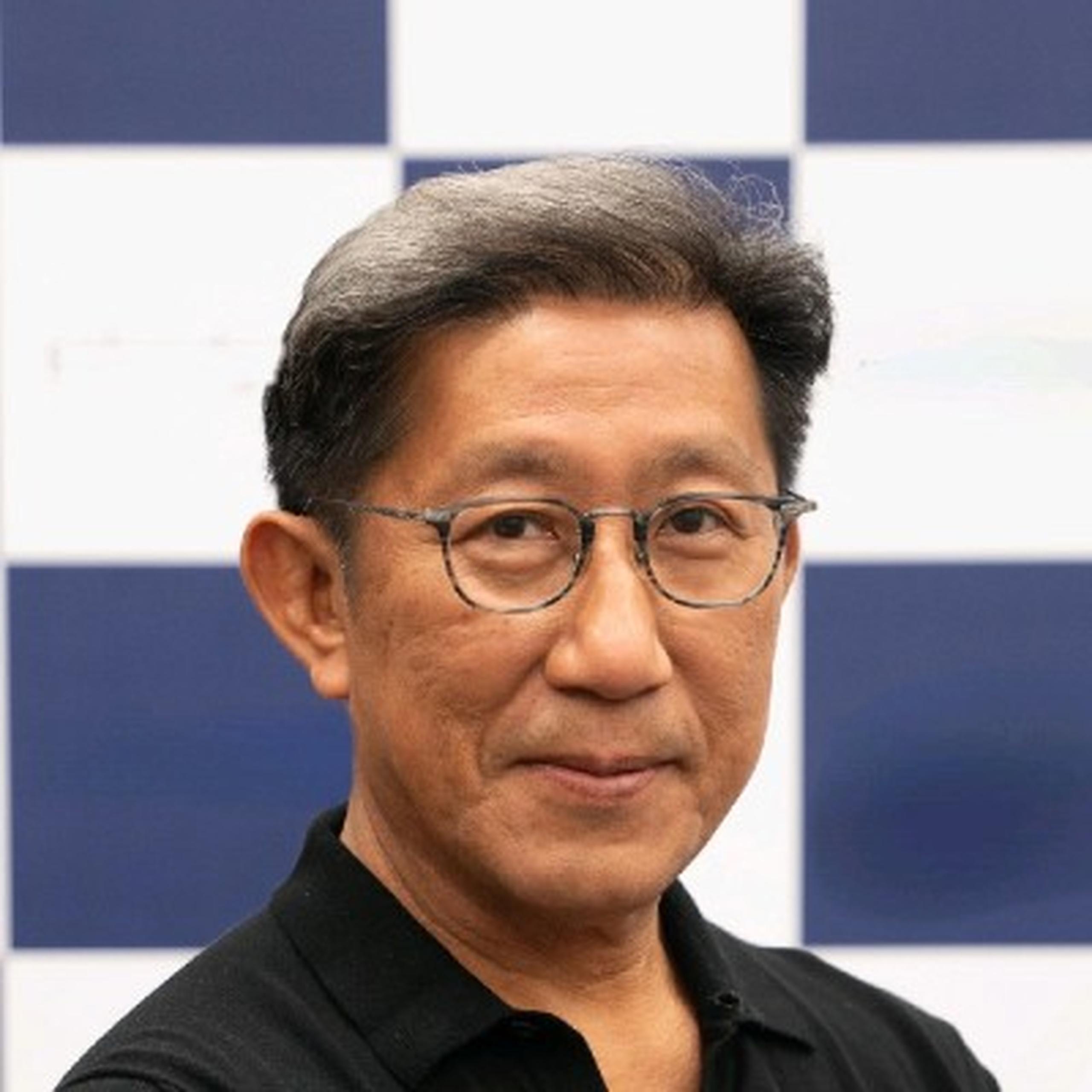 Man with glasses and gray hair, wearing a black shirt, poses in front of a blue and white checkered background.