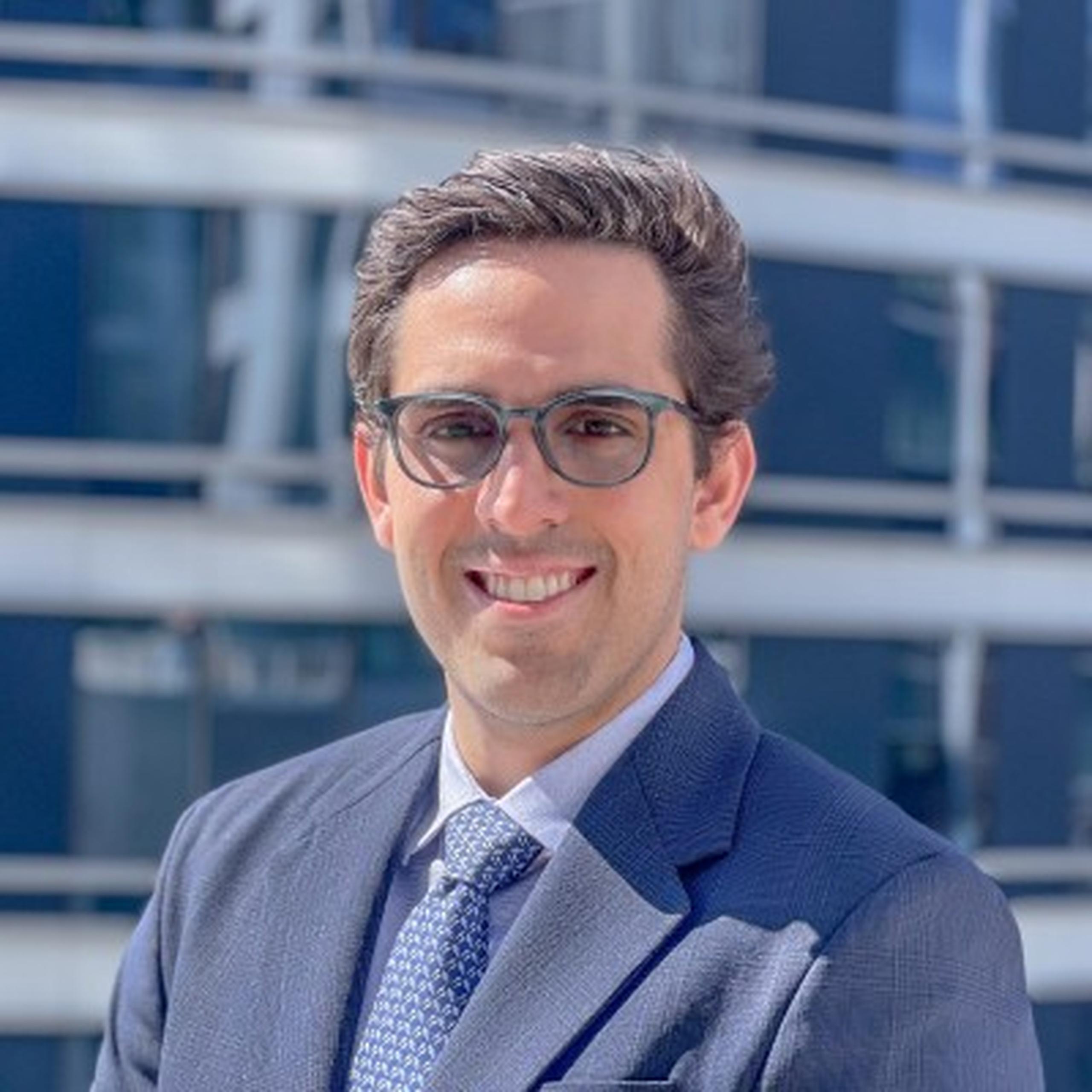 A person wearing glasses and a suit smiles outdoors with a modern building in the background.