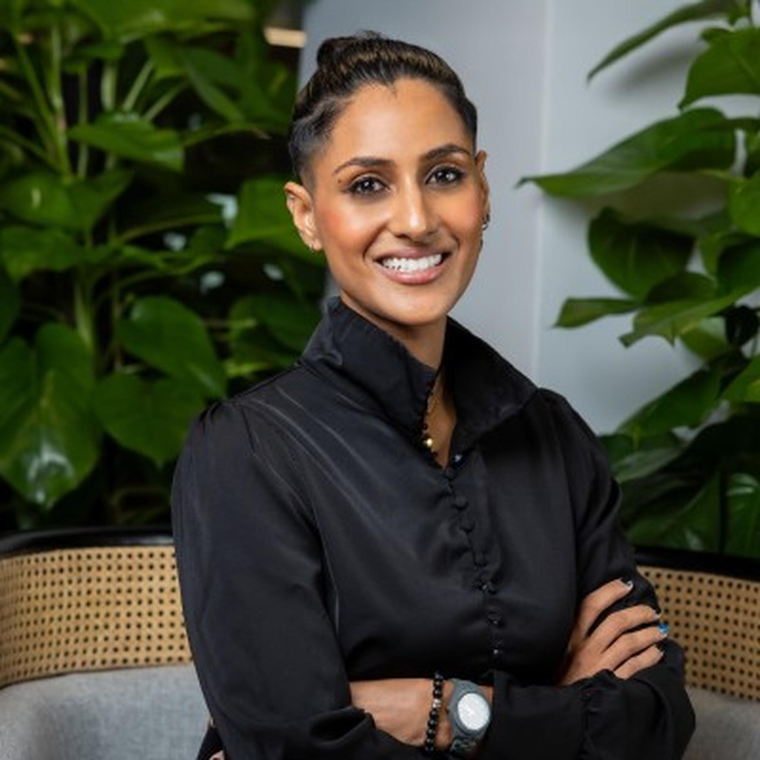 Smiling person in a black shirt with arms crossed, seated in front of lush green plants indoors.