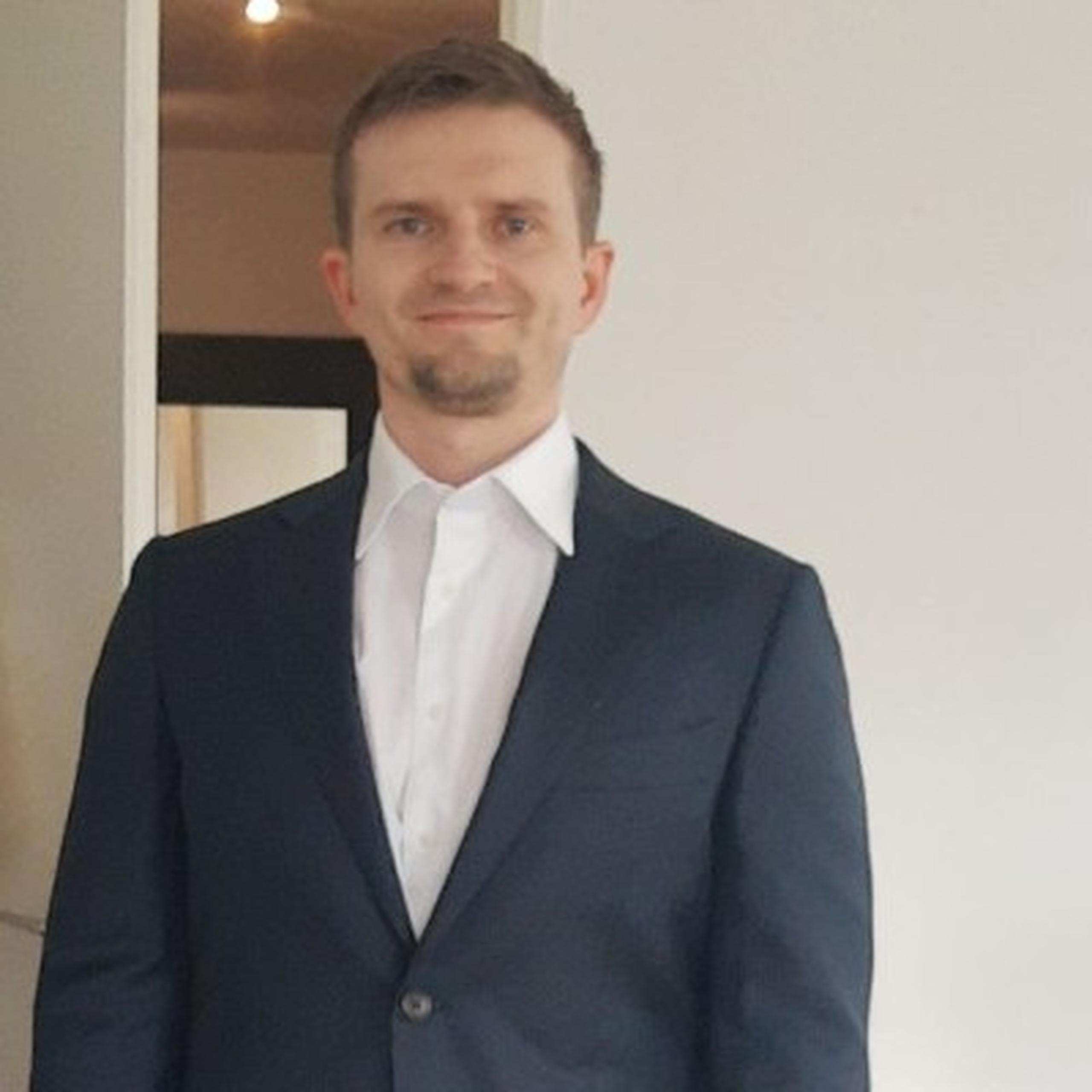 A man in a dark suit and white shirt, standing indoors against a plain wall, smiling slightly.