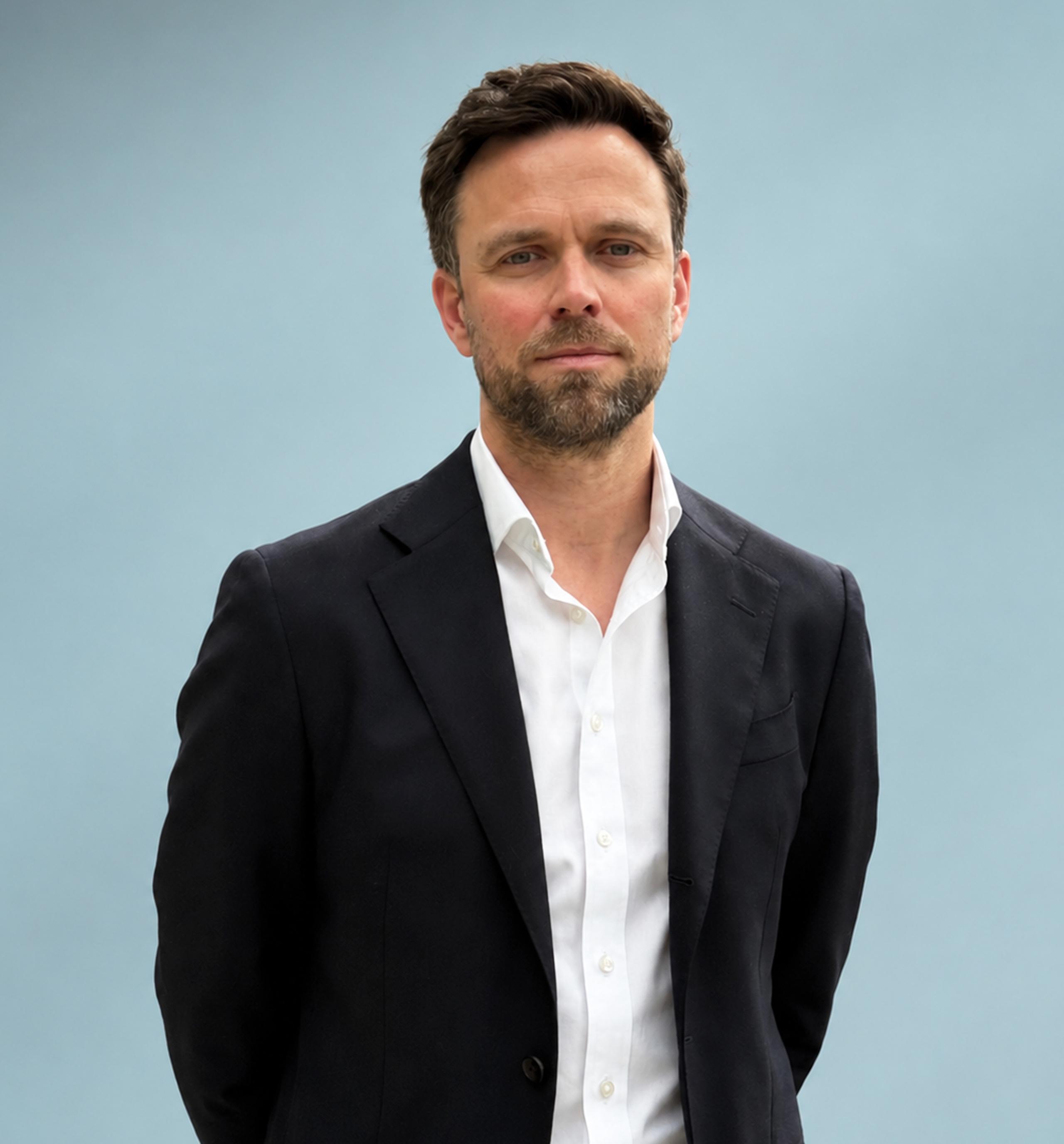 Man in a dark suit and white shirt standing against a light blue background, looking at the camera with a neutral expression.