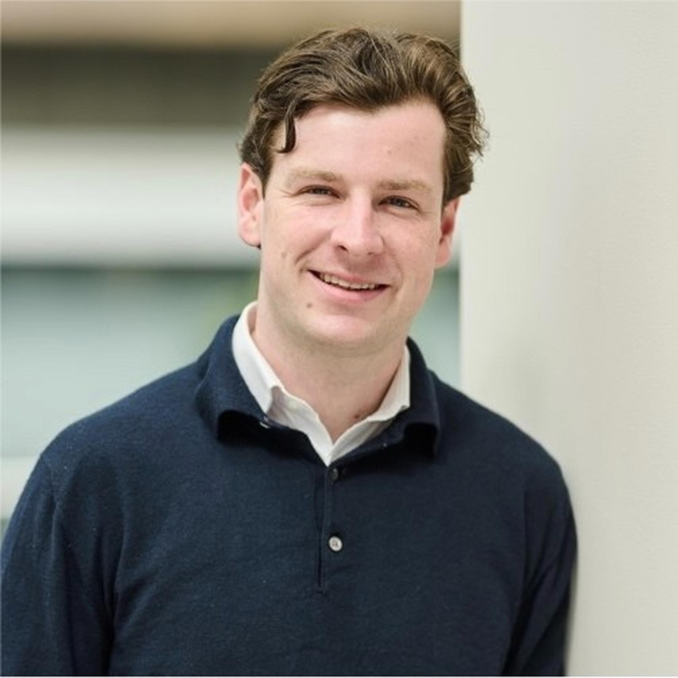 Smiling man with wavy brown hair in a navy sweater and white shirt, standing against a light-colored wall.